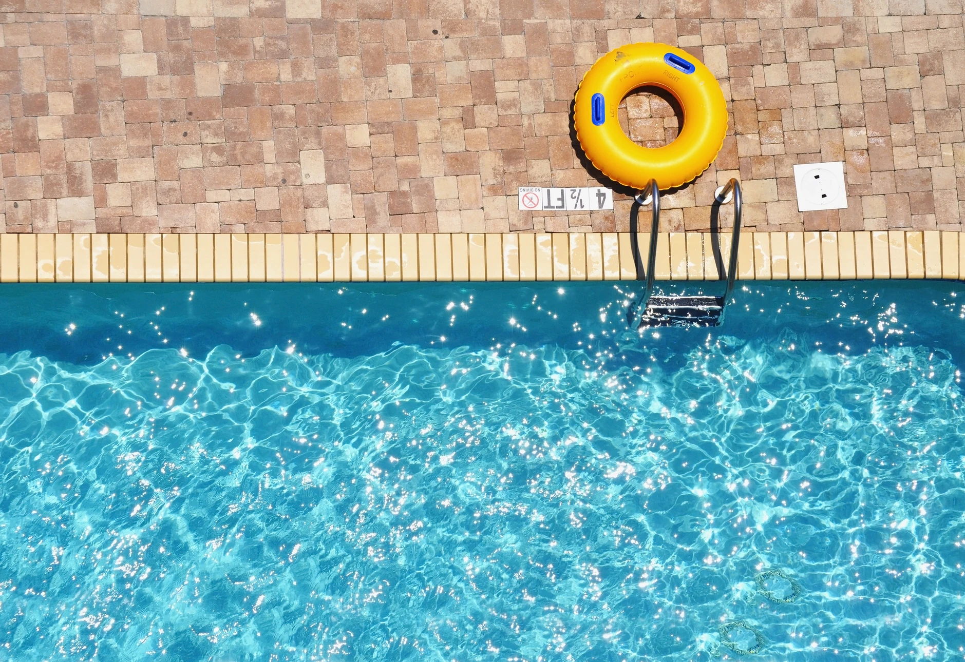 Piscine extérieure avec eau cristalline turquoise, margelles en pierre naturelle beige, escalier d'accès métallique, bouée de sauvetage jaune suspendue