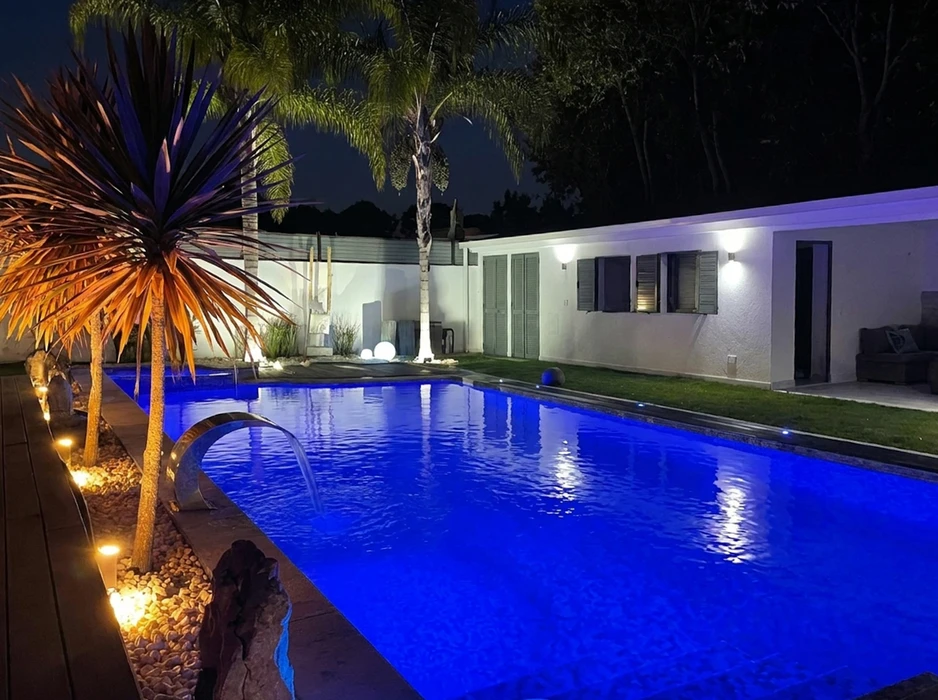 Piscine rectangulaire extérieure illuminée de nuit, eau bleue éclairée par LED subaquatiques, entourée d'un jardin paysager avec palmiers et cordyline rouge